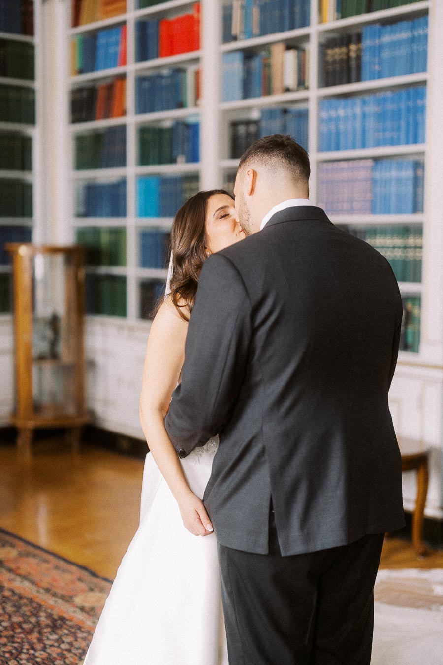 Bride and groom share a kiss in a library setting, surrounded by colorful books on shelves.