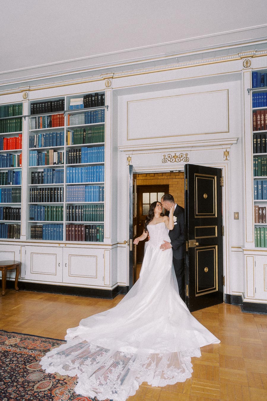 Bride and groom kissing in an elegant library with bookshelves, the bride wearing a long white gown with lace details.