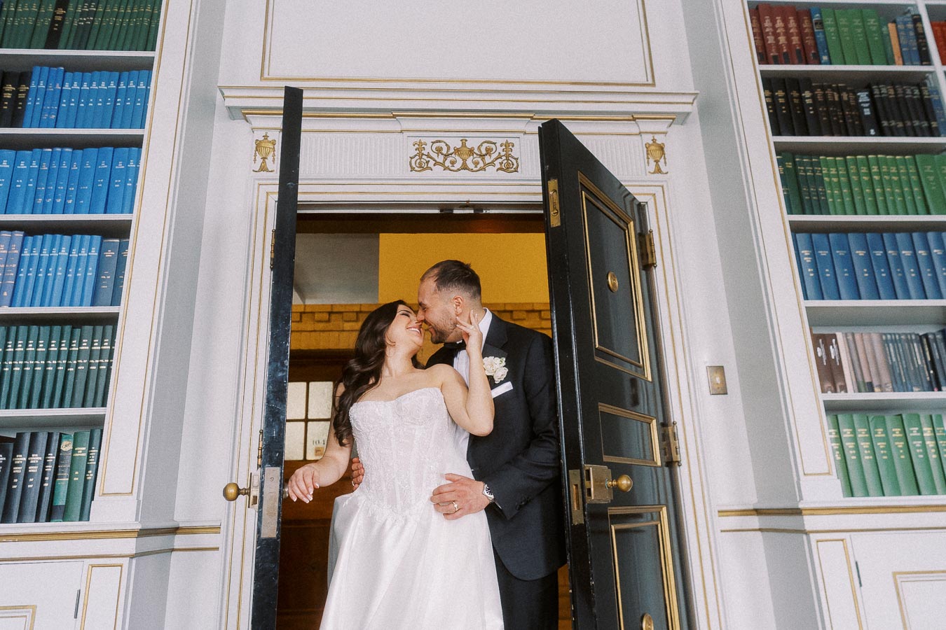 A newlywed couple sharing a joyful moment in a library setting, with the bride in a white dress and the groom in a suit, surrounded by bookshelves filled with colorful volumes.