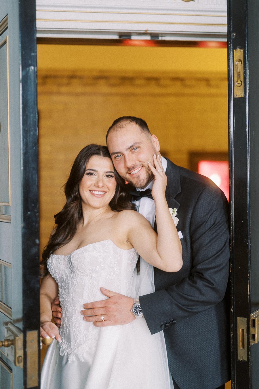 A happy bride and groom pose together at their wedding, smiling warmly while standing in a doorway. The bride is wearing a white lace strapless gown, and the groom is dressed in a dark suit with a white shirt and bowtie.