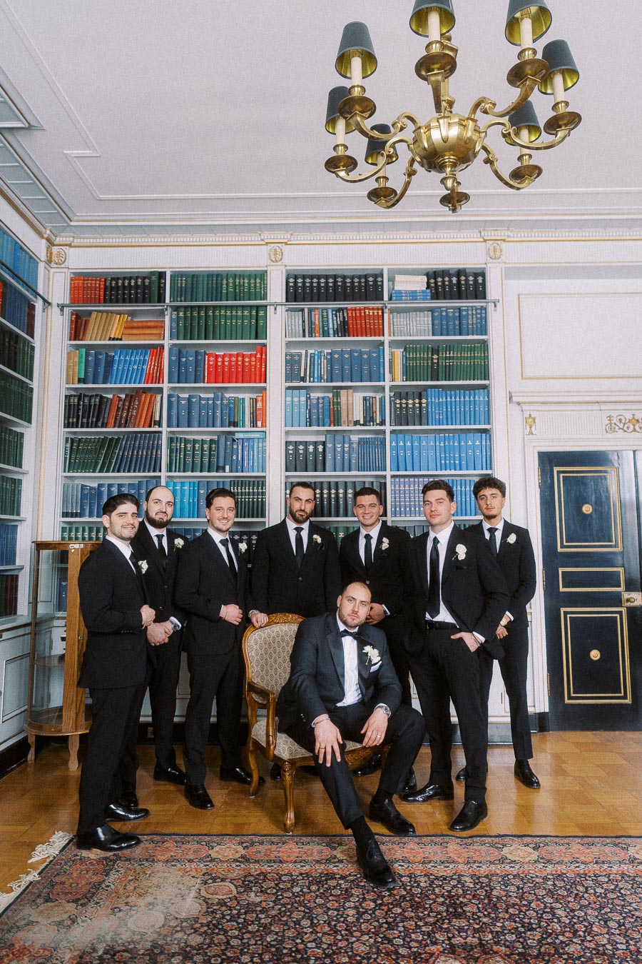 Groom and groomsmen in formal black suits posing in a sophisticated library setting with ornate bookshelves and chandelier, highlighting a classic wedding theme.