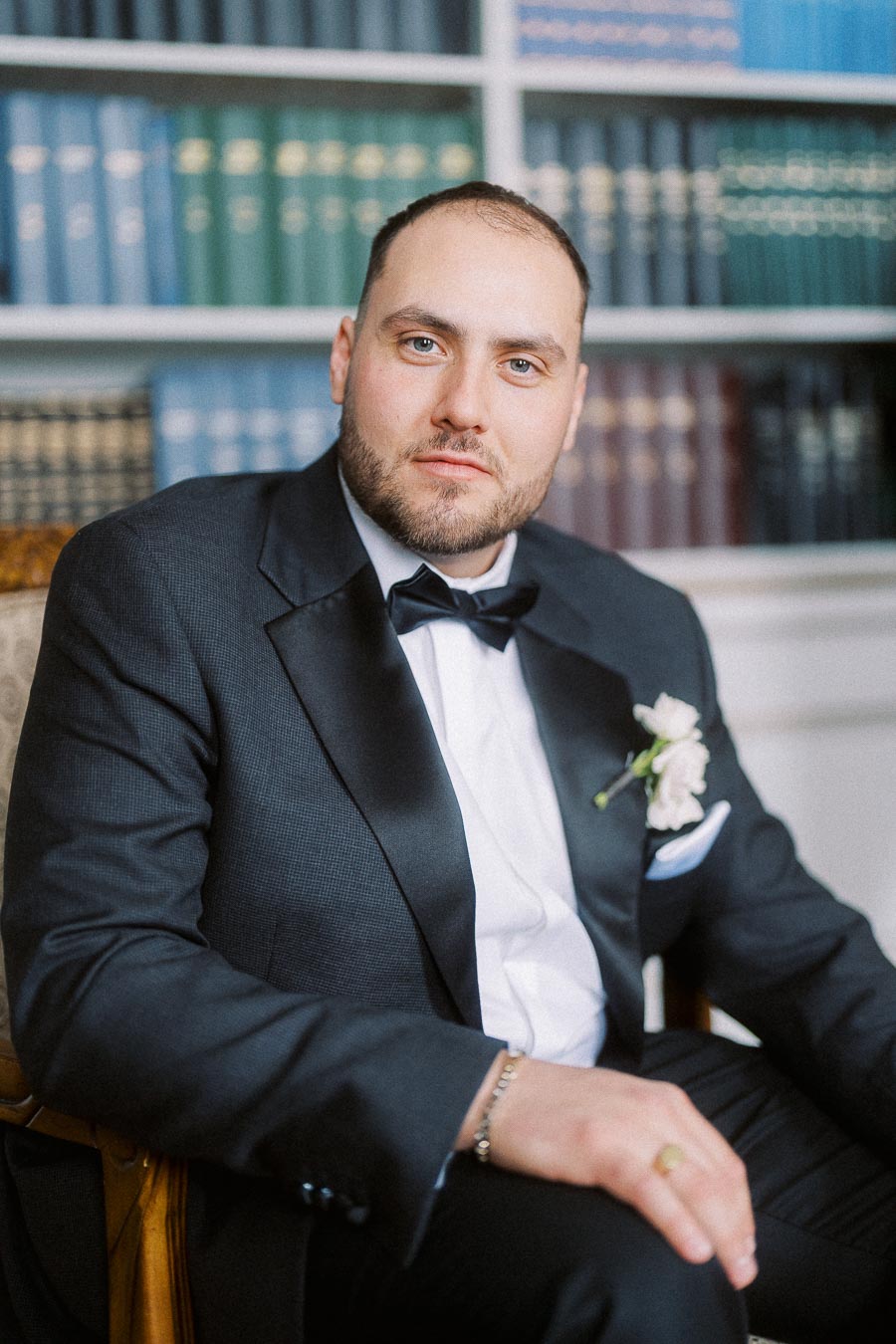 Well-dressed man in a tuxedo sitting in a library setting with elegant bookshelves in the background.