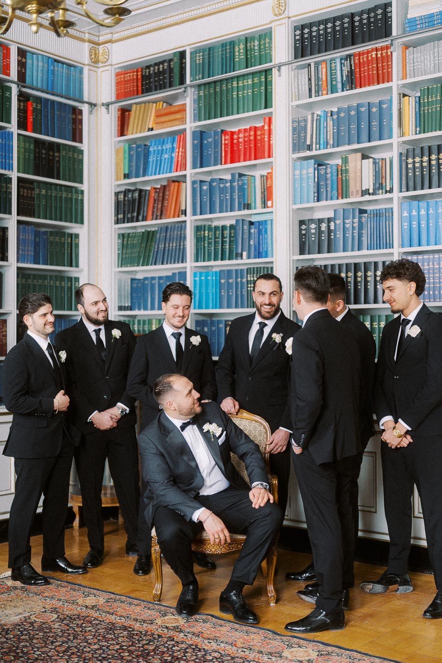Groom and groomsmen in elegant suits posing together in a stylish library setting with colorful bookcases in the background.