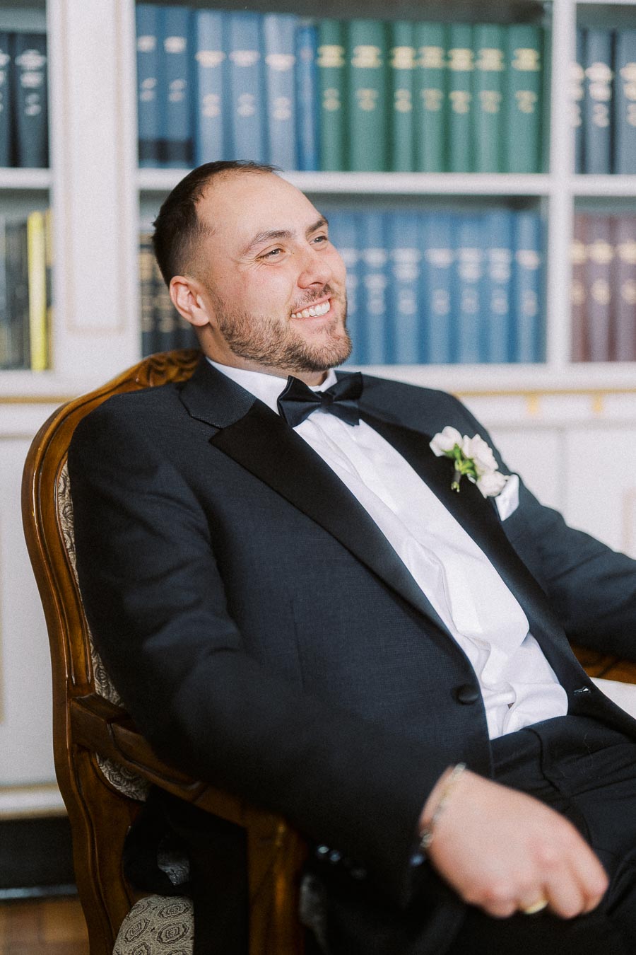 A man in a formal black tuxedo with a white shirt and bow tie sitting in a chair, smiling, with a library shelf filled with colorful books in the background.