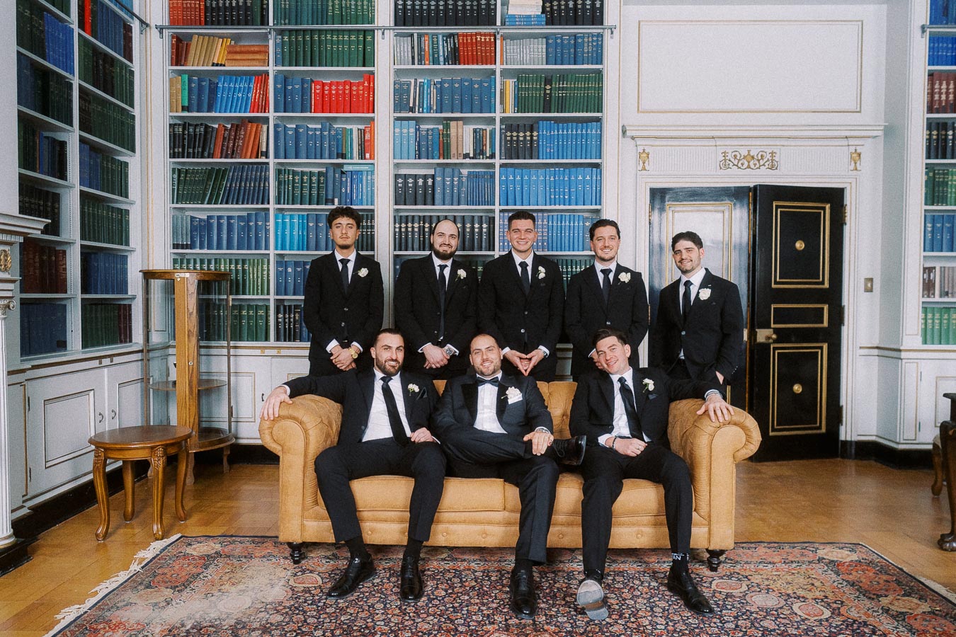 Group of eight groomsmen in black suits posing in a library with colorful bookshelves, two seated on a brown sofa and six standing behind, showcasing a formal wedding attire setting.
