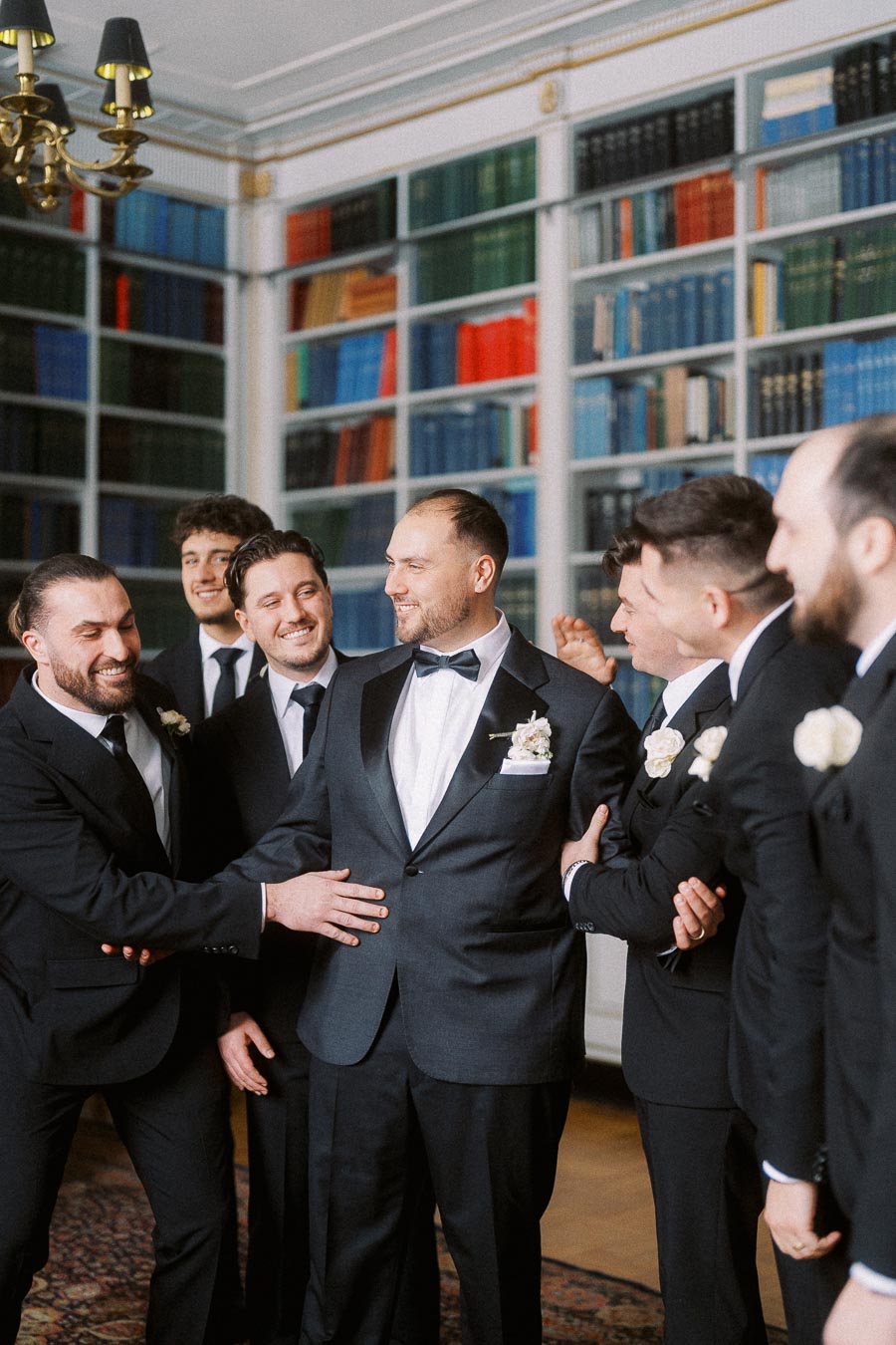 Groom in a black tuxedo surrounded by groomsmen in suits, laughing and celebrating in a library with colorful bookshelves.