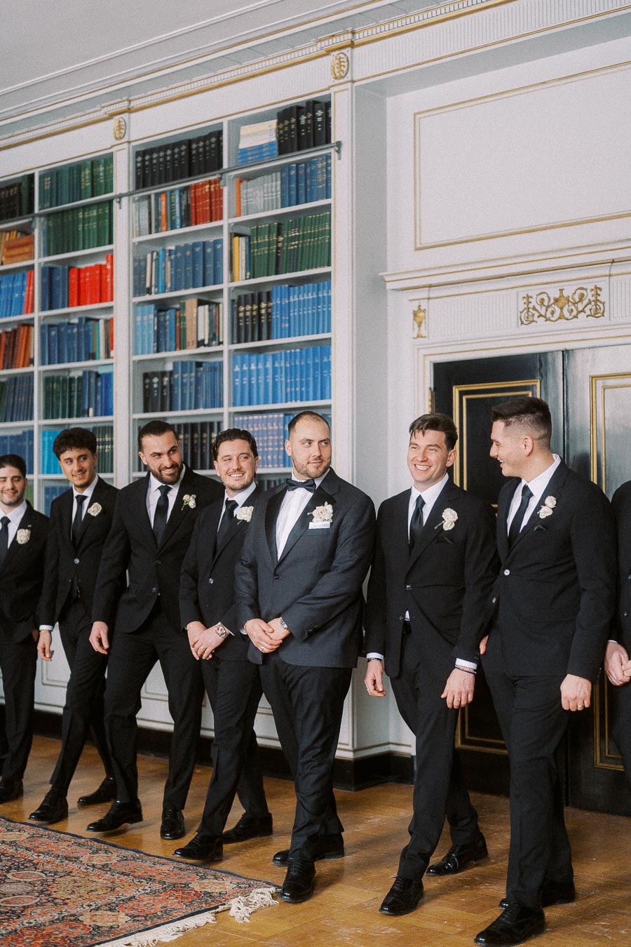 A group of groomsmen in formal suits and boutonnieres, standing together in a library setting with colorful bookshelves in the background.
