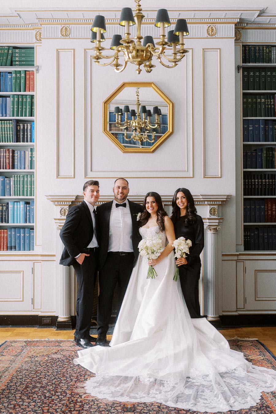 A wedding party posing in an elegant library setting. A bride in a flowing white gown stands with her bridal party, holding white bouquets. They are surrounded by an ornate interior with a chandelier and bookshelves filled with colorful books.