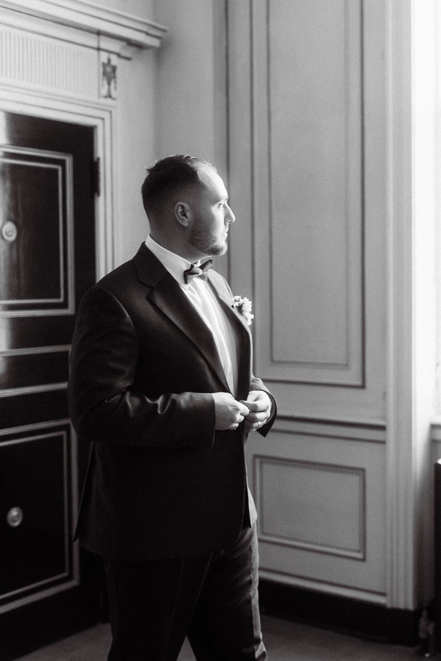 Black and white photo of a groom in a suit and bow tie, standing indoors and gazing out a window, captured in a classic and elegant setting.