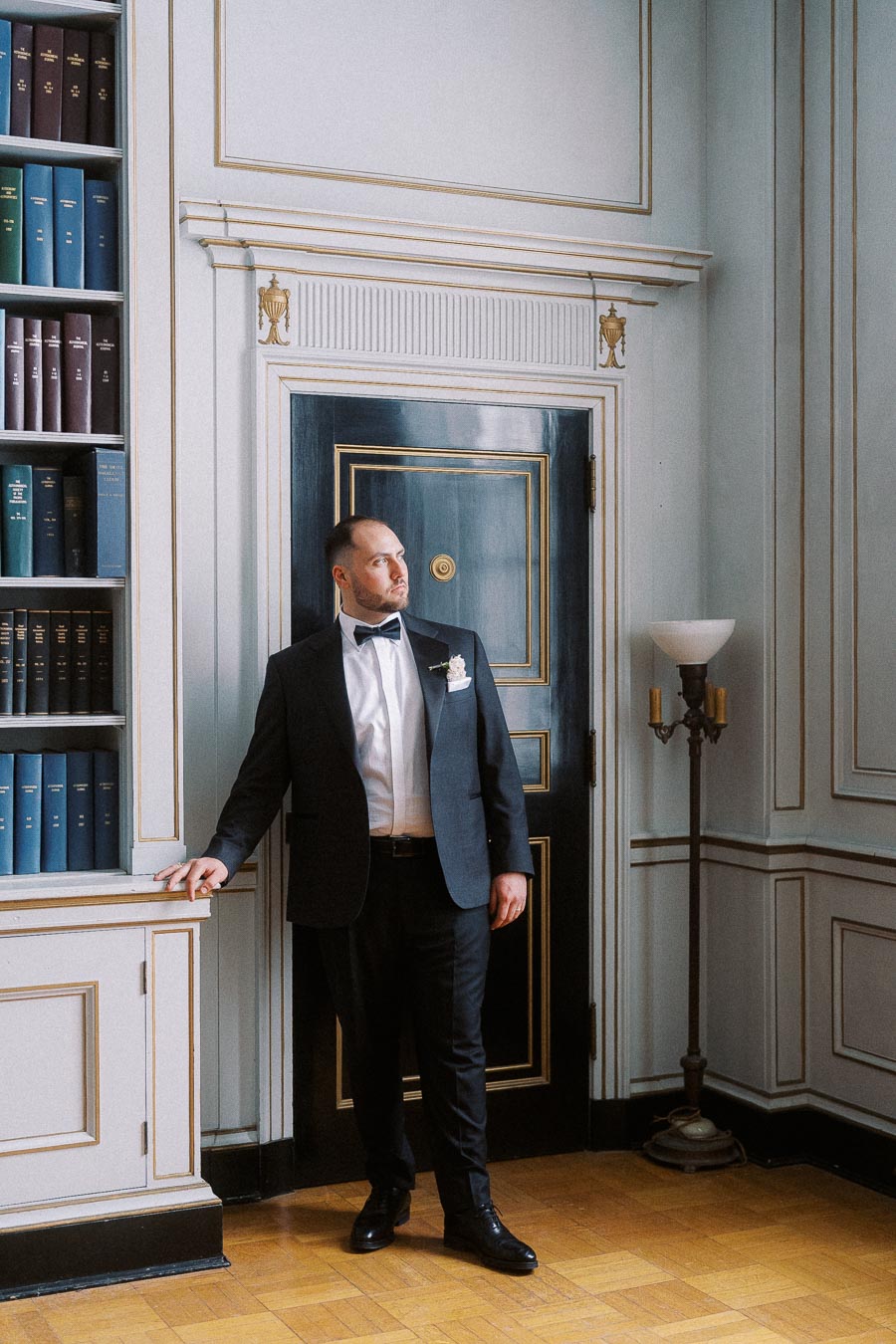A well-dressed man in a formal suit stands in an elegant room with classic decor, featuring a bookshelf filled with colorful books and ornate paneling.