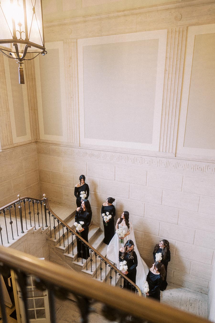 Bridal party in elegant black dresses with white bouquets descending a grand staircase in a classic, ornate building.
