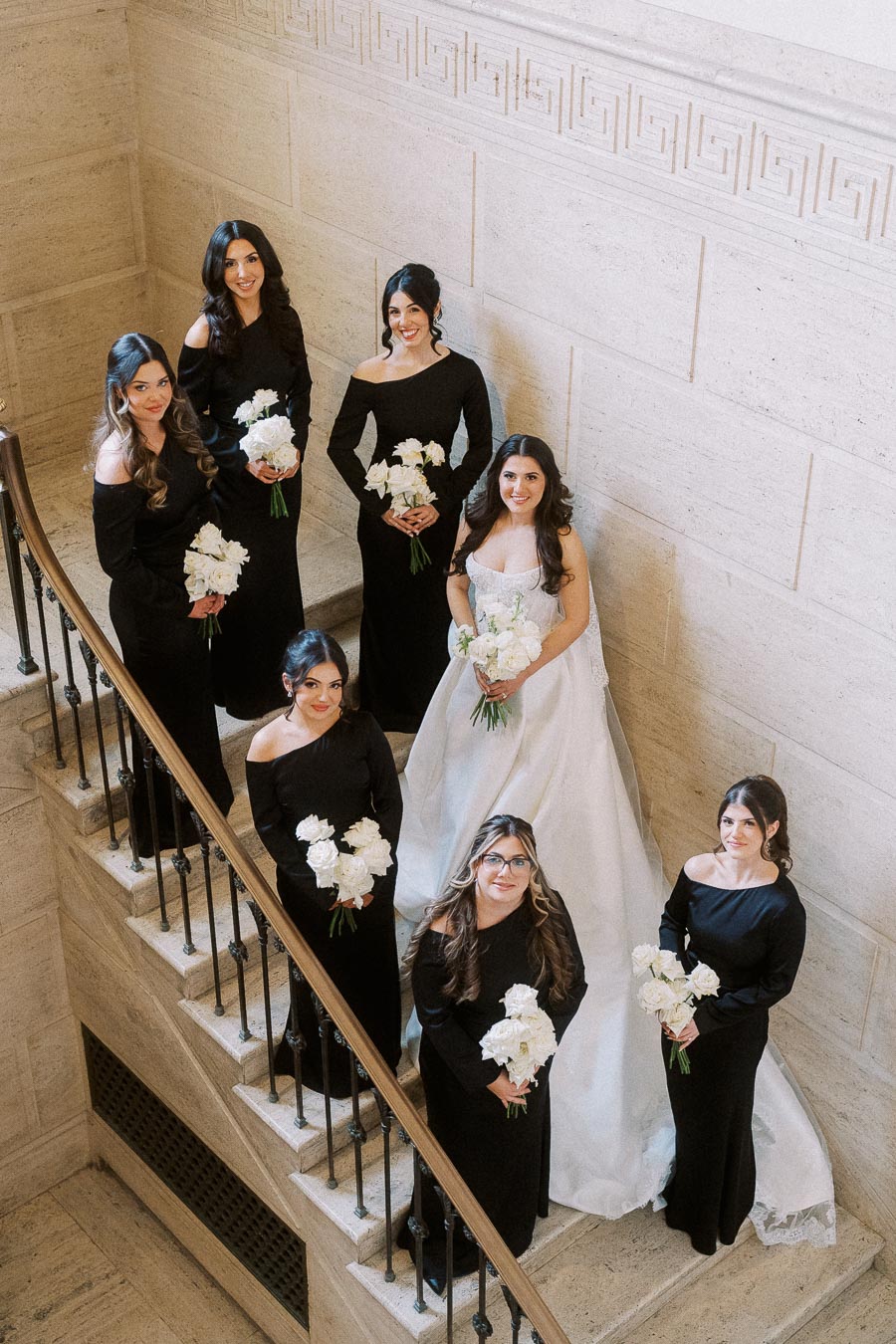 Bridal party poses on a grand staircase, with the bride in a white gown surrounded by bridesmaids in elegant black dresses holding white bouquets.