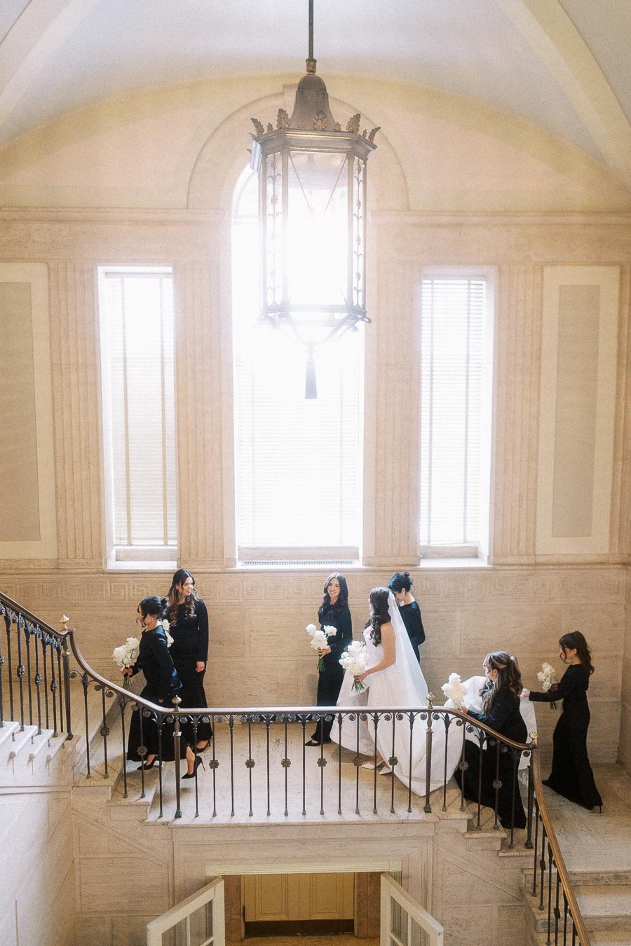 Bridal party ascending elegant staircase in a historic venue, featuring a bride in a white wedding gown and bridesmaids in black dresses holding bouquets, with natural light streaming through large windows.