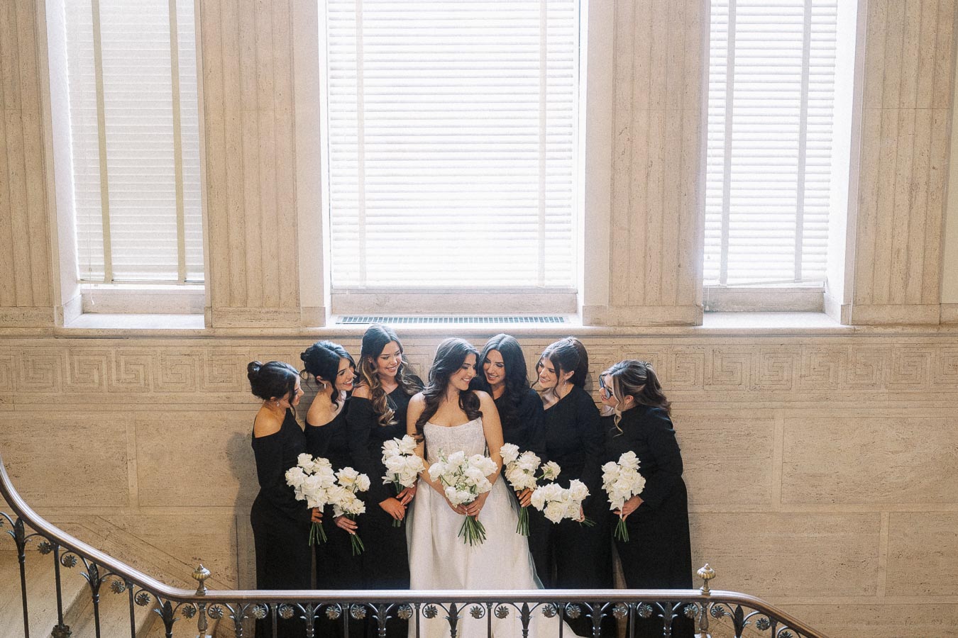 A bride in a white gown stands smiling with her bridesmaids, all dressed in black, holding white bouquets in an elegant stone interior with large windows.