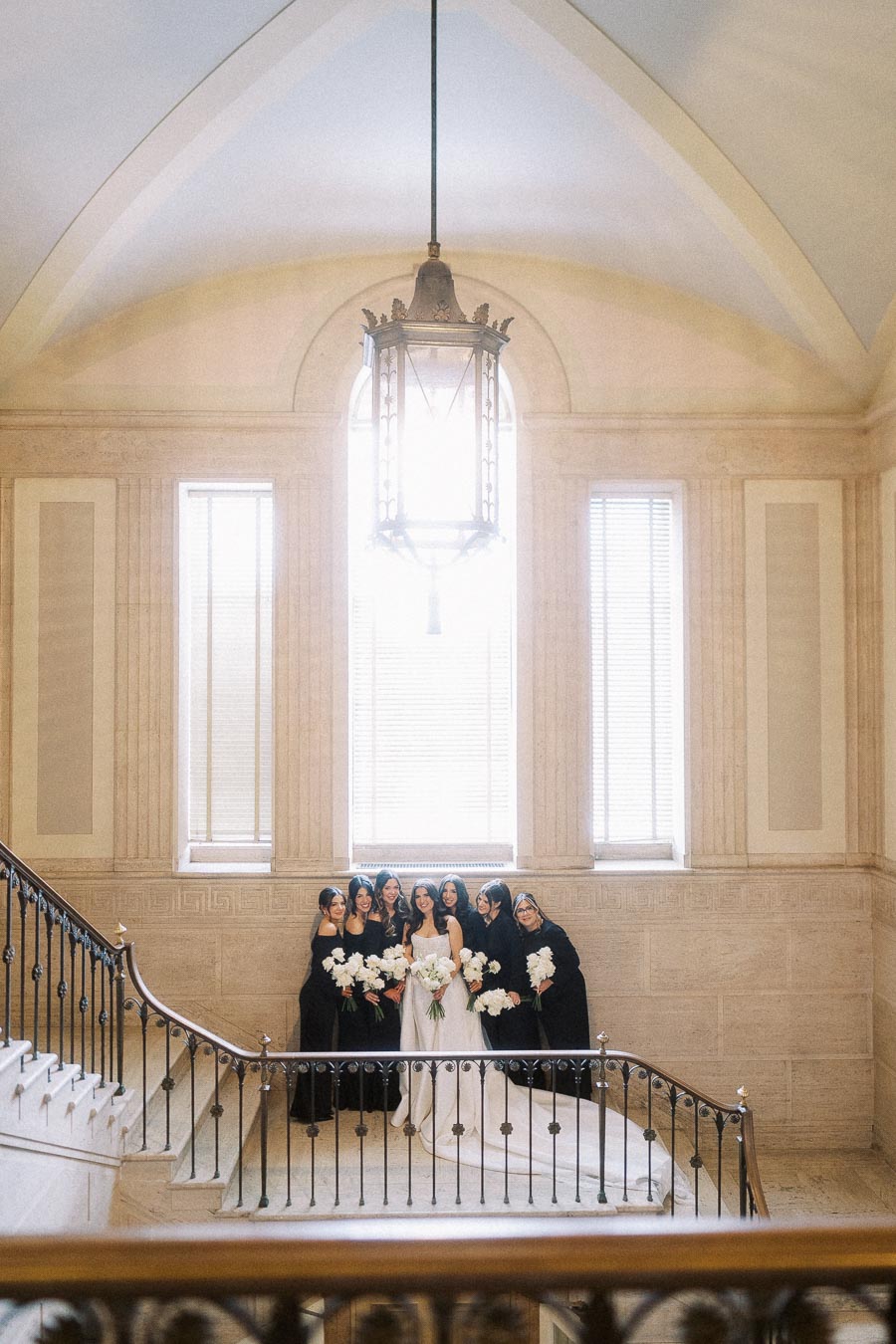 Bridal party posing on elegant staircase with large windows and ornate lighting, featuring a bride in a white gown surrounded by bridesmaids in black dresses holding white bouquets.