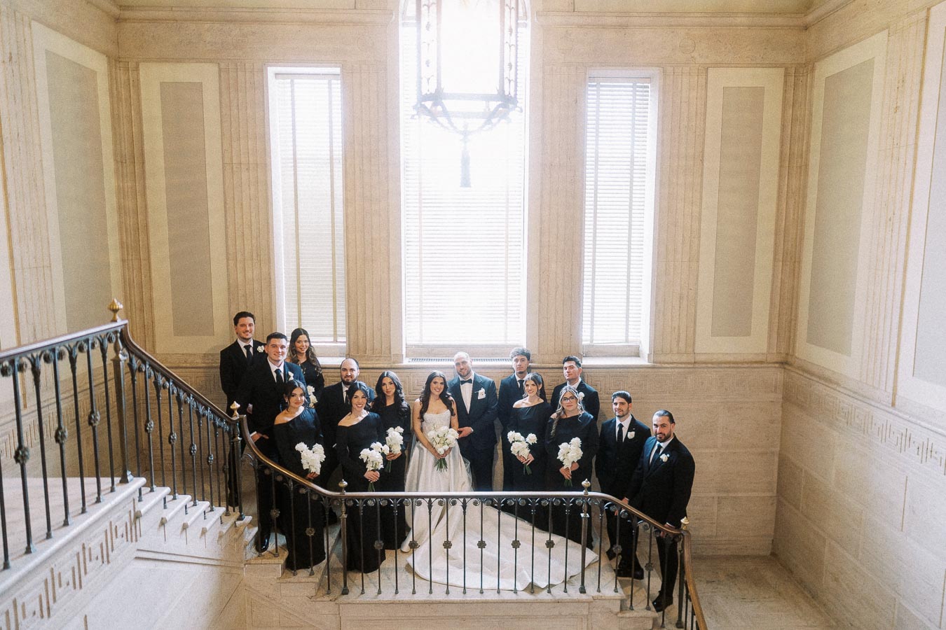 Elegant wedding party posing on a grand staircase, featuring a bride in a white gown, groom in a tuxedo, and attendants in black formal attire holding bouquets, set against an ornate interior backdrop.