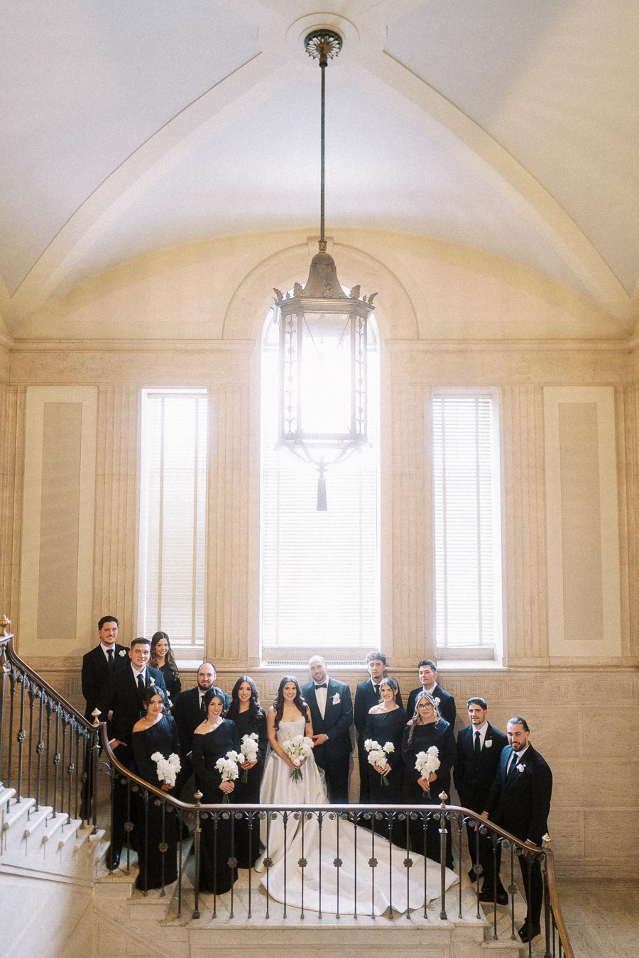 A wedding party poses on a grand staircase in an elegant venue, with the bride and groom surrounded by bridesmaids in matching black dresses and groomsmen in black suits, under a large ornate chandelier against a backdrop of tall windows.