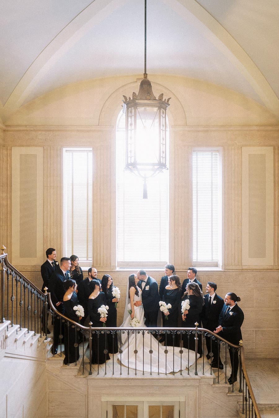 Elegant wedding party gathered on a grand staircase with a bride and groom surrounded by bridesmaids and groomsmen, all dressed in formal attire, inside a beautifully lit architectural setting.