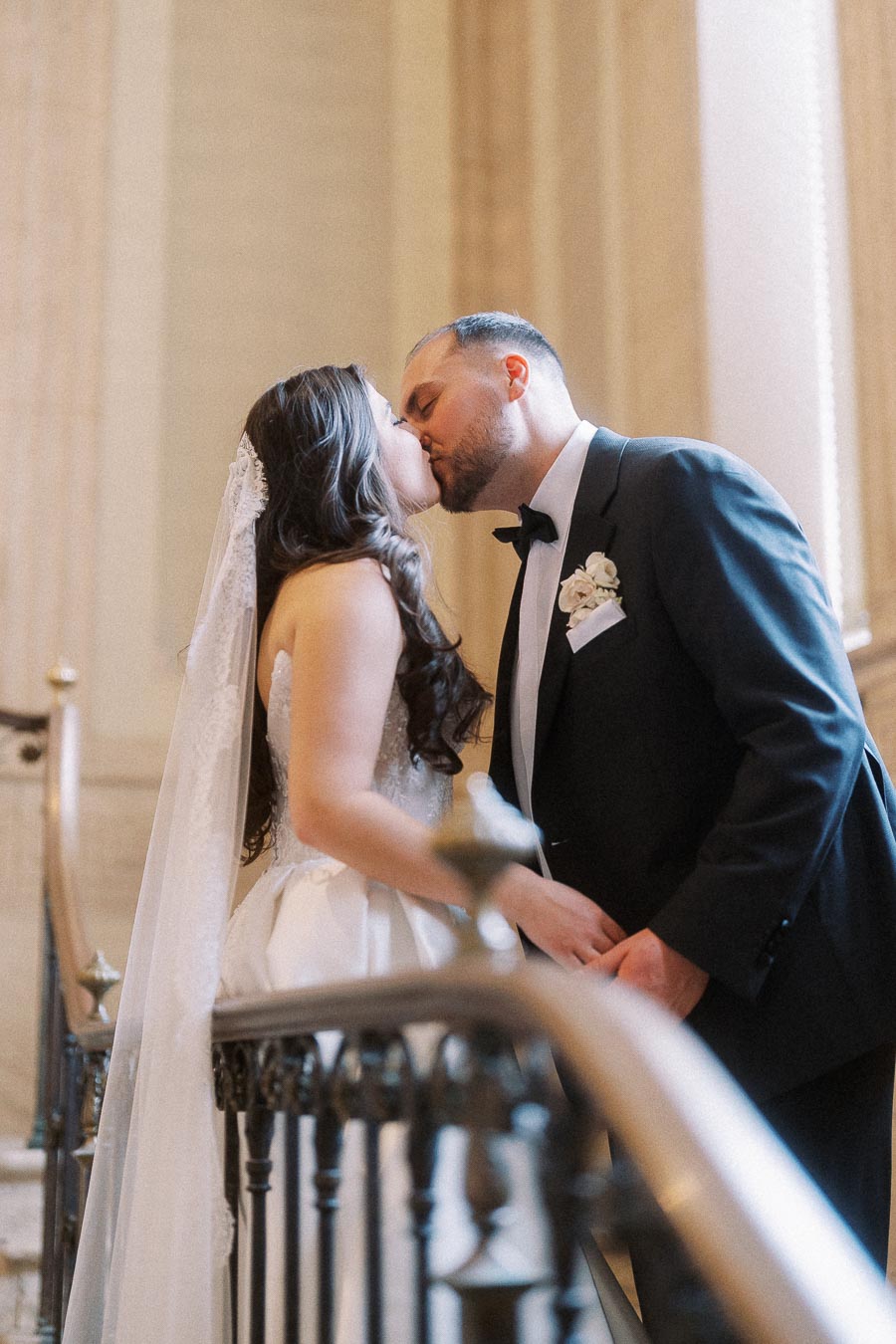 Bride and groom sharing a kiss on a grand staircase, with the bride wearing an elegant white gown and veil and the groom in a black tuxedo, capturing a romantic wedding moment.