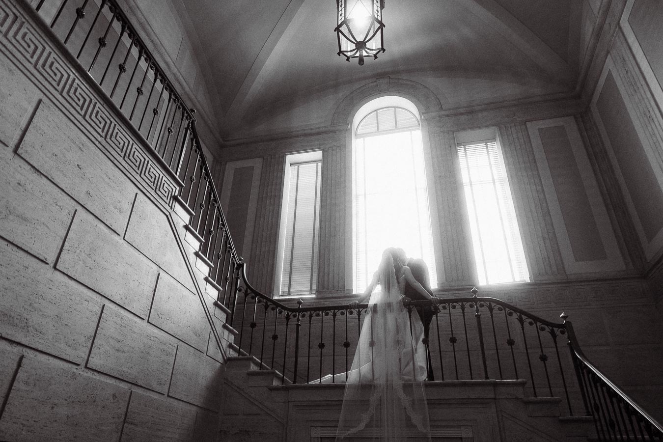 A black and white image of a couple embracing on an elegant staircase with a large window, ideal for wedding photography inspiration.