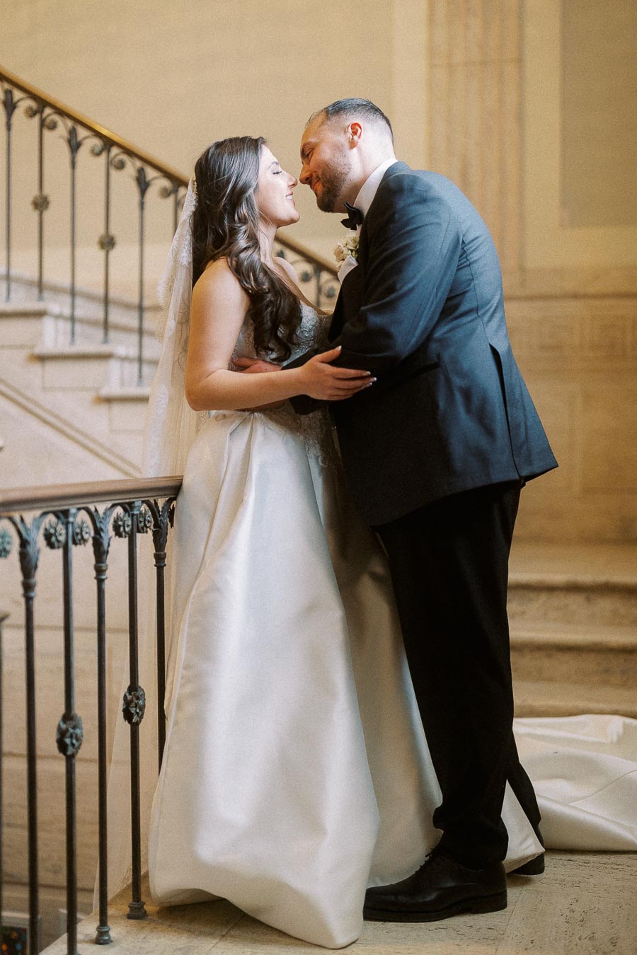 Bride and groom sharing a romantic moment on a grand staircase, she in a flowing white wedding gown, and he in a classic black suit.