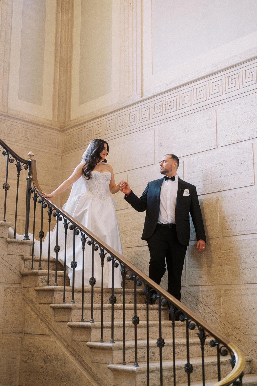 Bride and groom descending an elegant staircase in a historic building, showcasing a romantic moment on their wedding day. The bride is in a classic white gown and the groom in a sharp black suit.