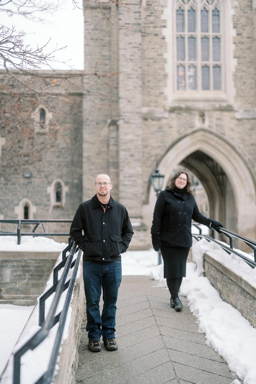 A man and woman walking on a snow-covered pathway near a historic stone building with arched windows, dressed in winter clothing.