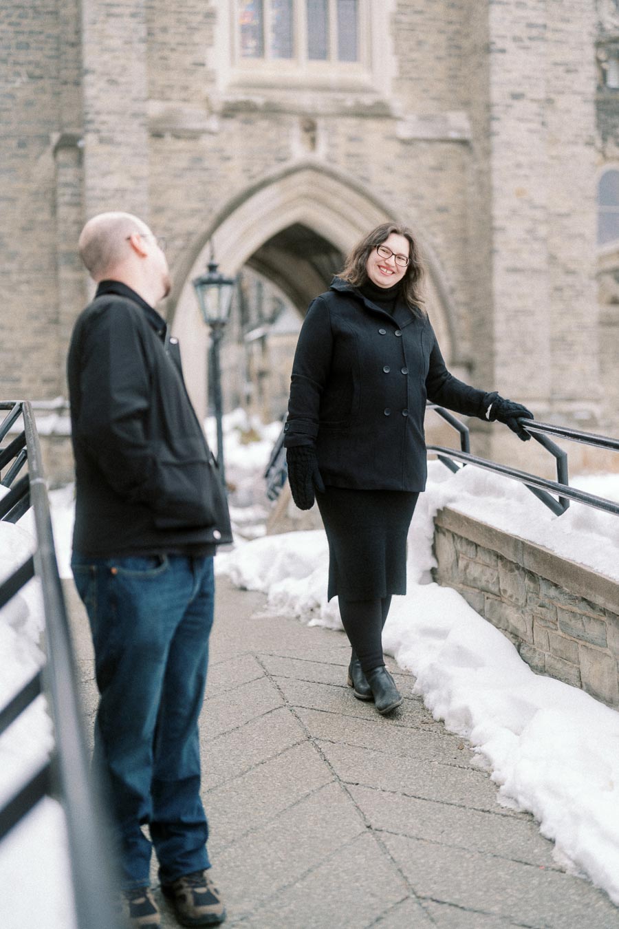 A woman smiling on a snowy walkway in front of a historic stone building, wearing a black coat and gloves, while a man in casual clothing stands nearby.