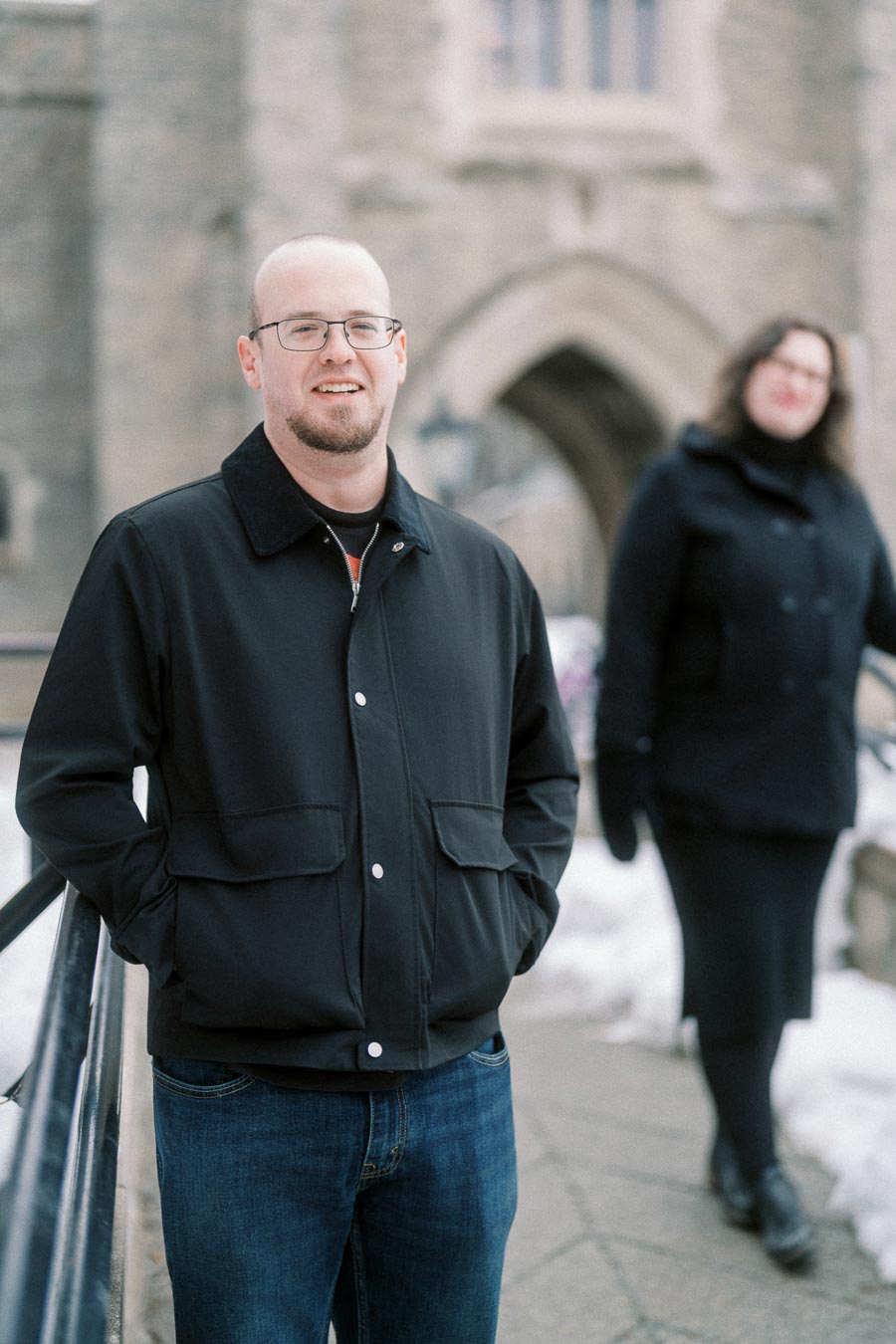 Man in a black jacket standing outdoors on a snowy pathway, with a woman in a dark outfit blurred in the background near a stone building.