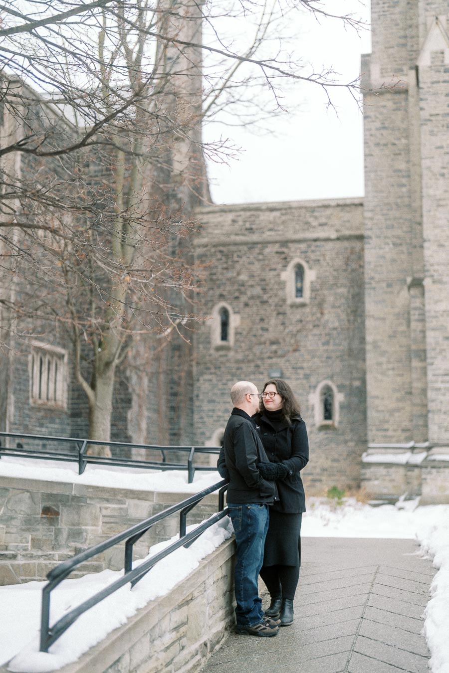 A couple dressed in winter clothing stand close together on a snowy pathway, surrounded by a historic stone building and bare trees.