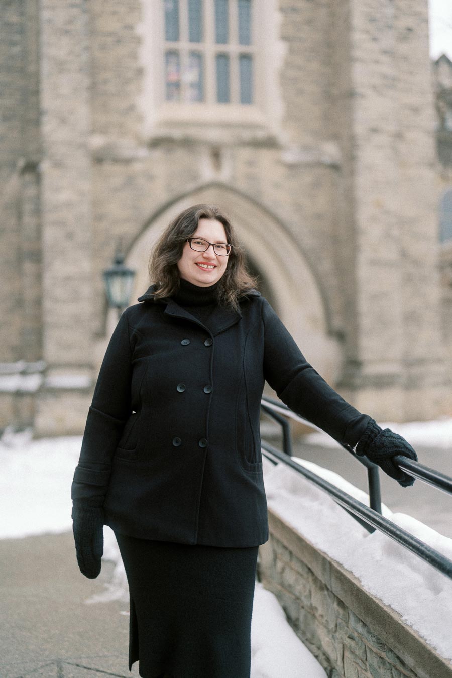 Woman in black coat smiling in front of a historic stone building during winter.
