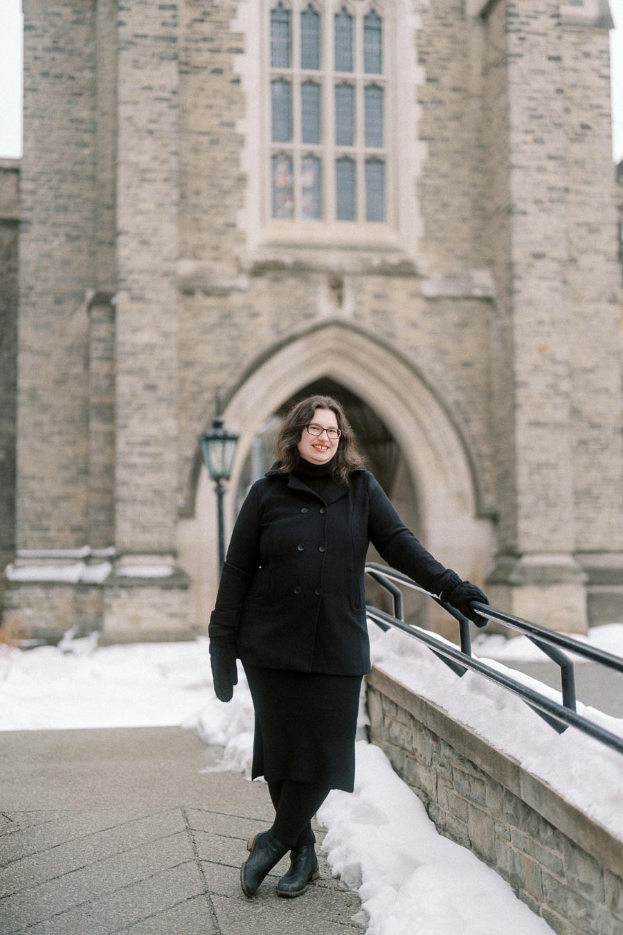 Person in winter clothing smiling and leaning on a railing in front of a historic stone building with snow on the ground.