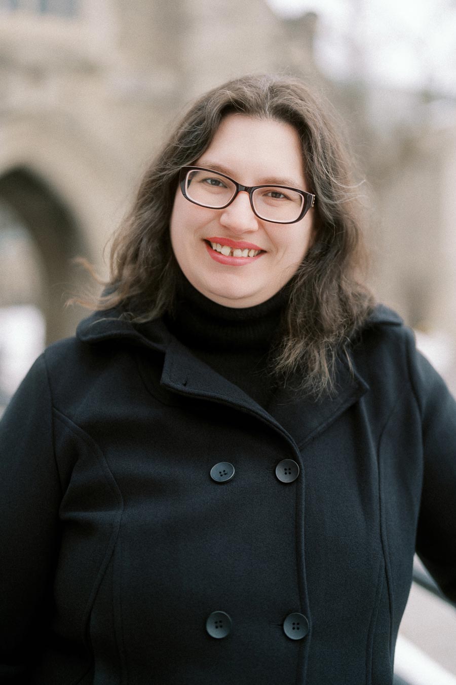 Portrait of a smiling woman in a black coat and glasses, standing outdoors with a blurred architectural background.