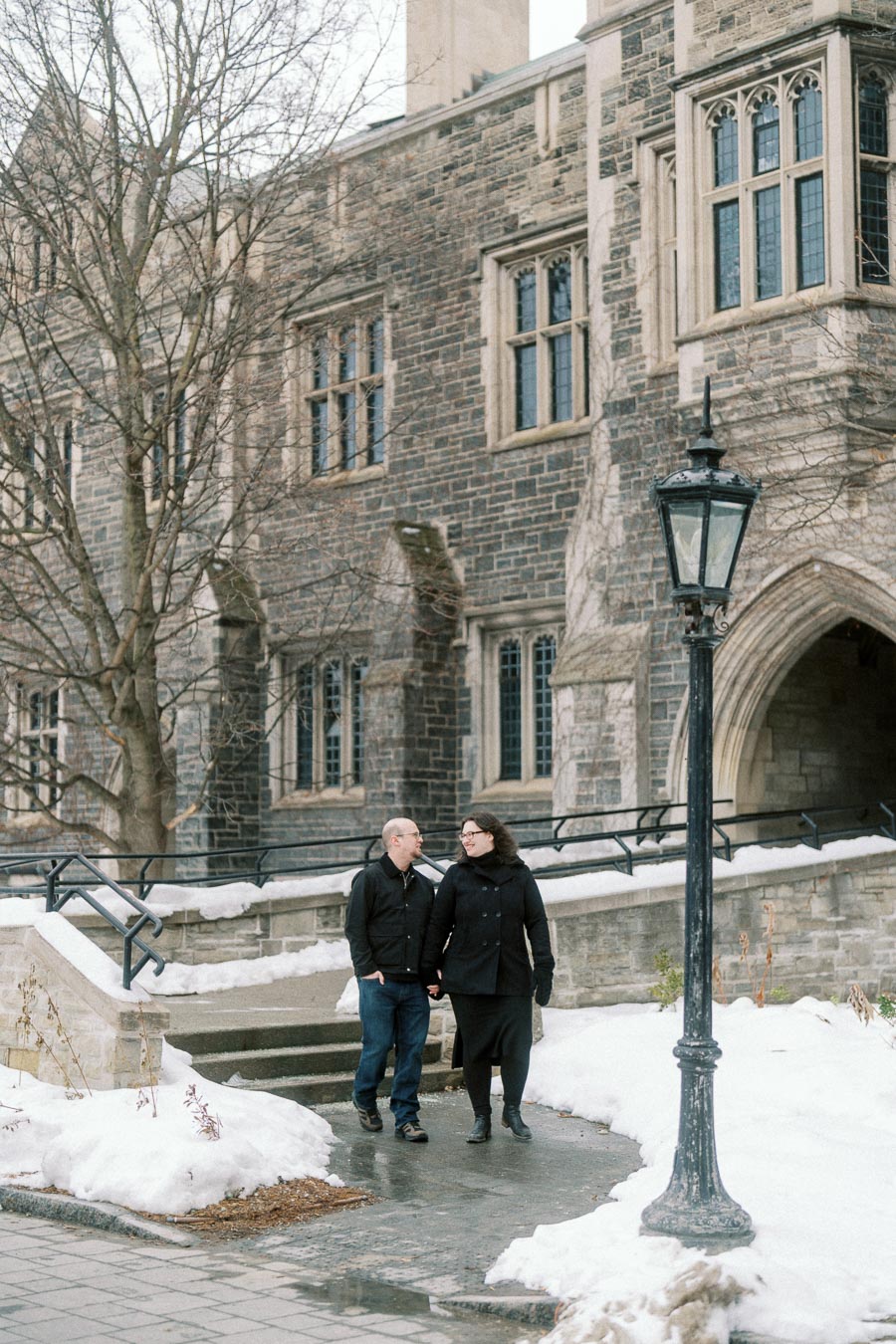 A couple walking hand in hand on a snowy path in front of a historic stone building with arched windows and a vintage streetlamp, capturing a serene winter moment.