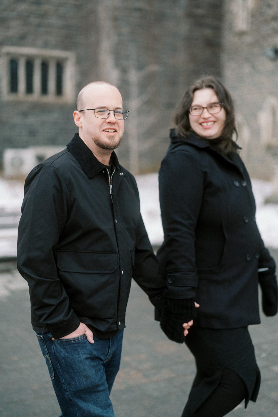 A couple in winter attire holding hands and walking outdoors in front of a stone building.