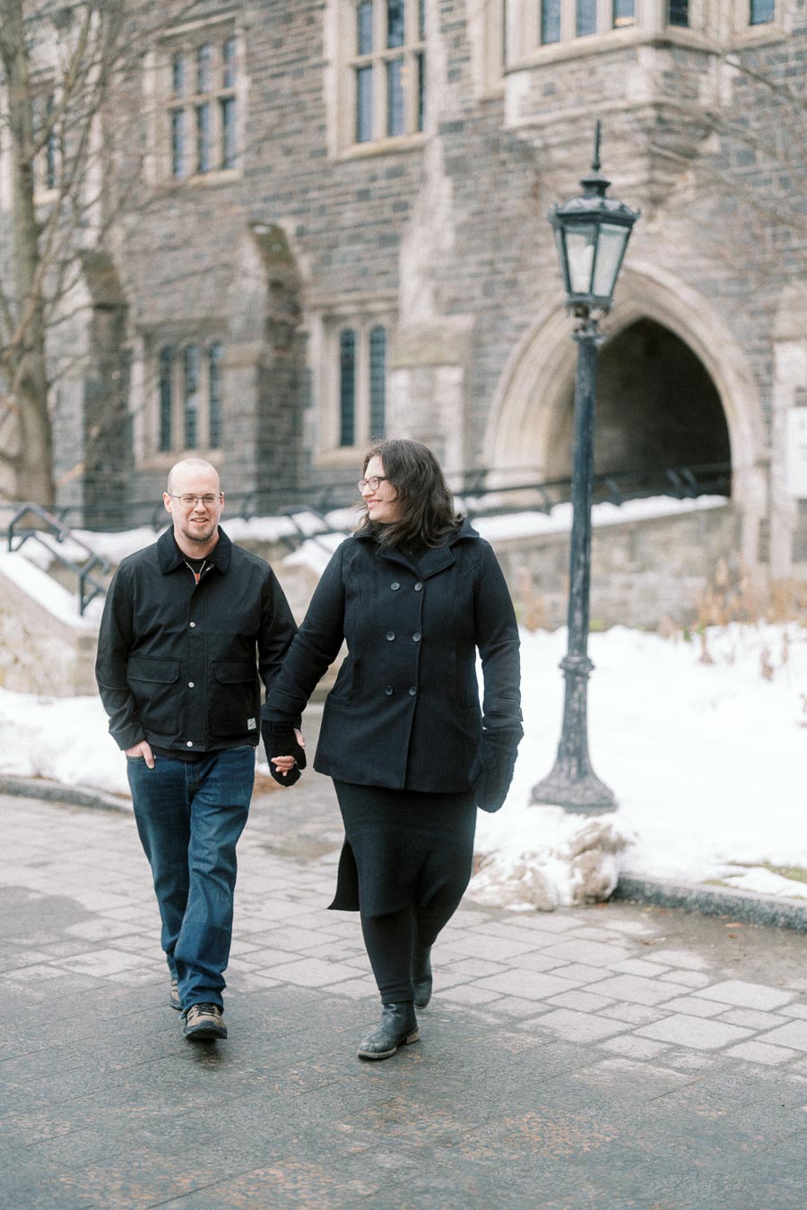 A couple holding hands and walking on a snowy pathway in front of a historical stone building with gothic architecture and a vintage street lamp.