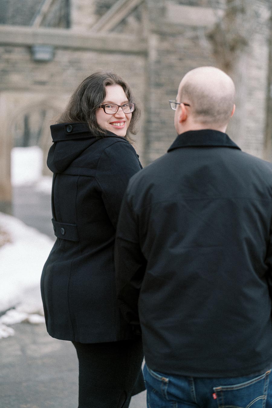 Happy woman in black coat smiling while looking over her shoulder at a man in a snowy setting with stone architecture in the background.
