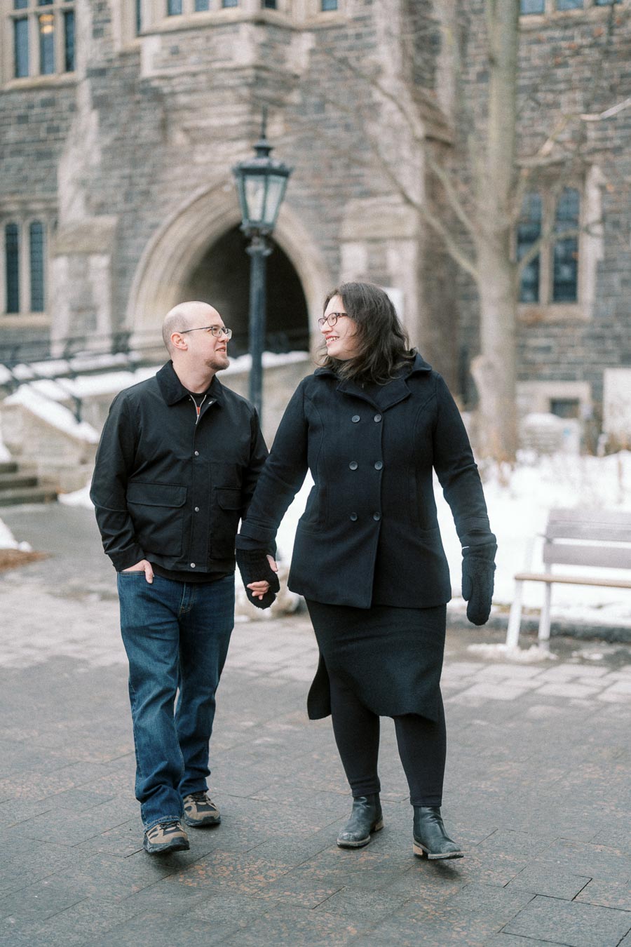 A couple in winter coats walking hand in hand outside a historic stone building with arched windows and a lamppost, surrounded by a snowy landscape.