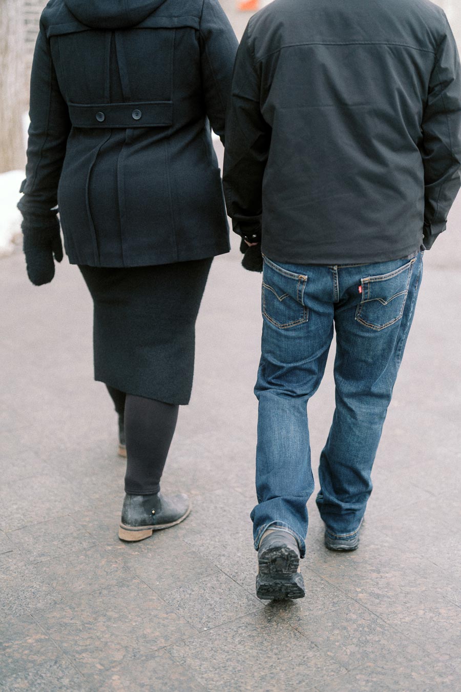 Rear view of a couple walking on a sidewalk in winter, wearing warm jackets and jeans, holding hands.