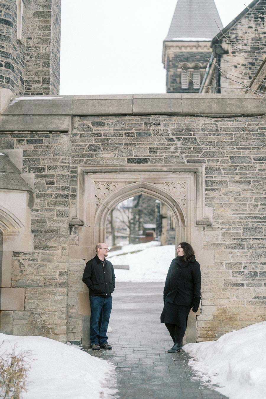 A man and woman stand facing each other under a stone archway at a historic building, surrounded by snow.