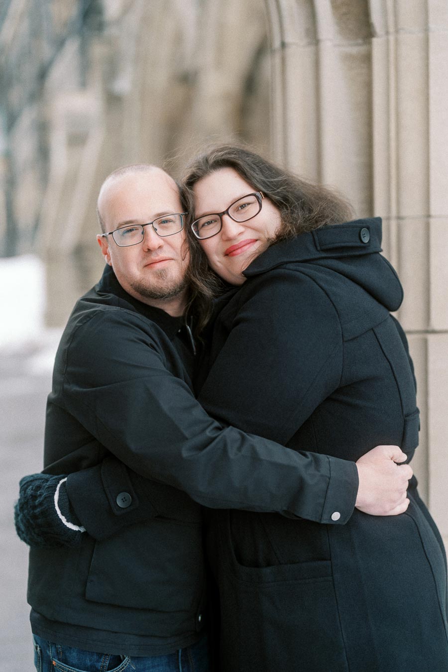 A couple wearing black coats embracing and smiling in front of a stone building.
