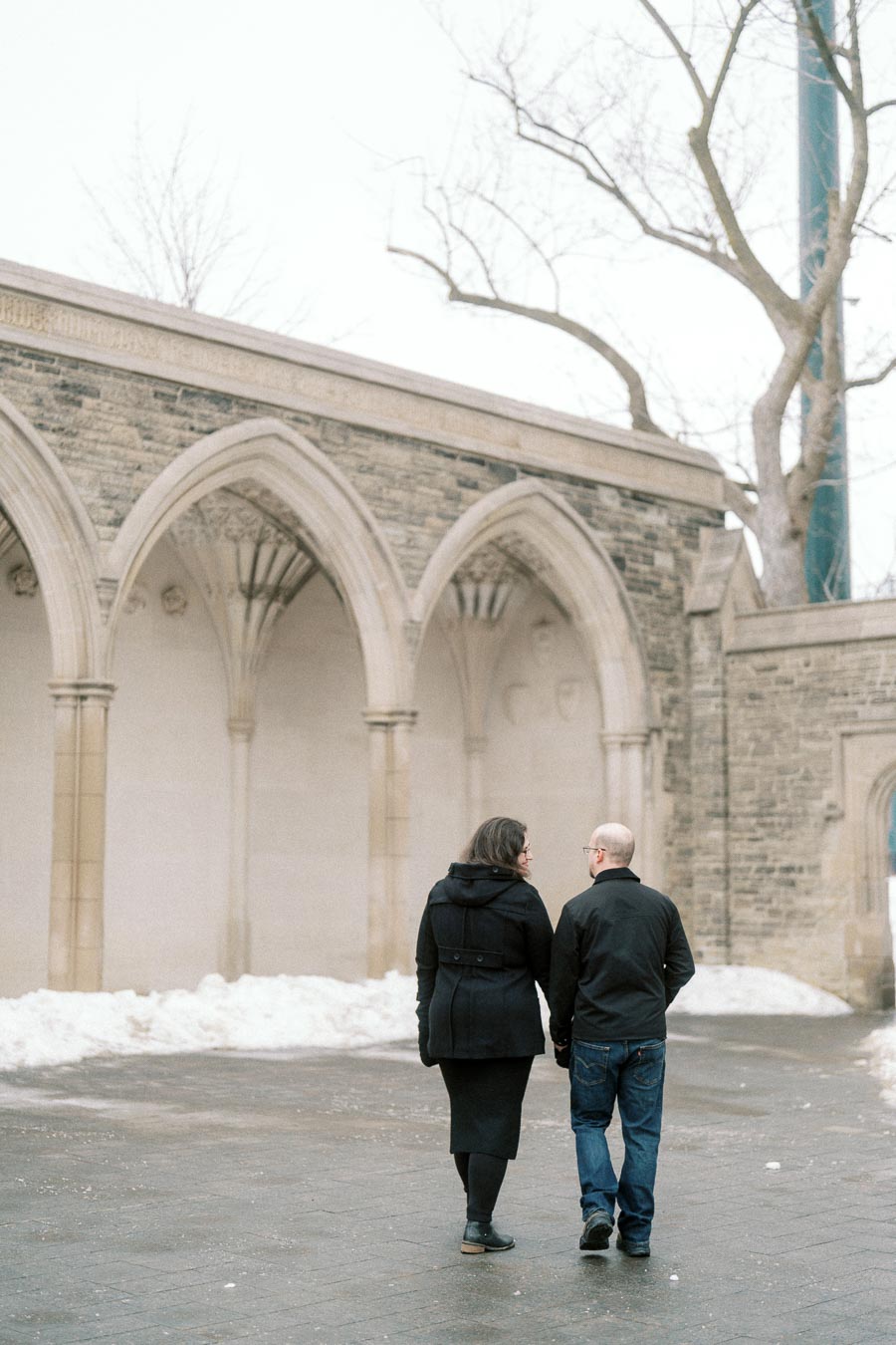 A couple walks hand in hand through a historic stone archway, surrounded by a light layer of snow, symbolizing romance and tranquility in a winter setting.