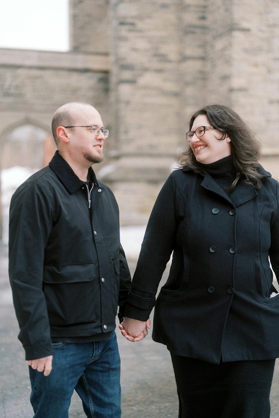 A couple holding hands and smiling at each other in front of a historic stone building, wearing winter coats.