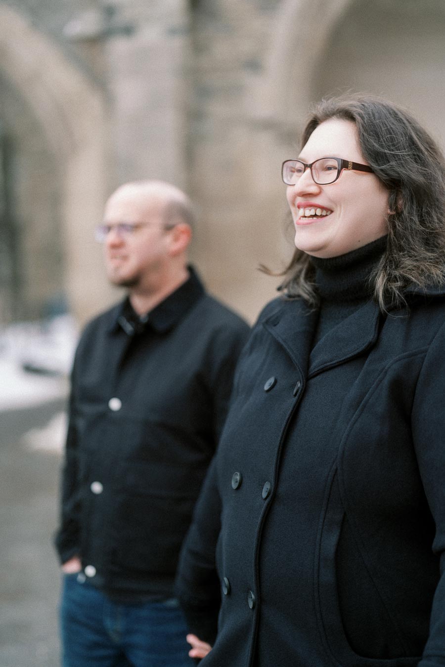 Two people standing outdoors wearing black coats, with one smiling and the background featuring blurred stone architecture.
