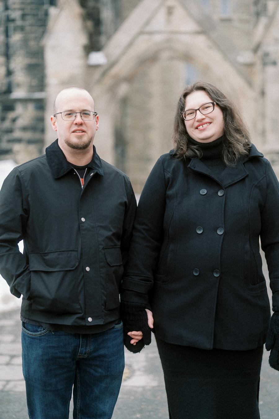 A couple poses in front of a historic stone building, both wearing dark coats and glasses, holding hands, and smiling warmly.