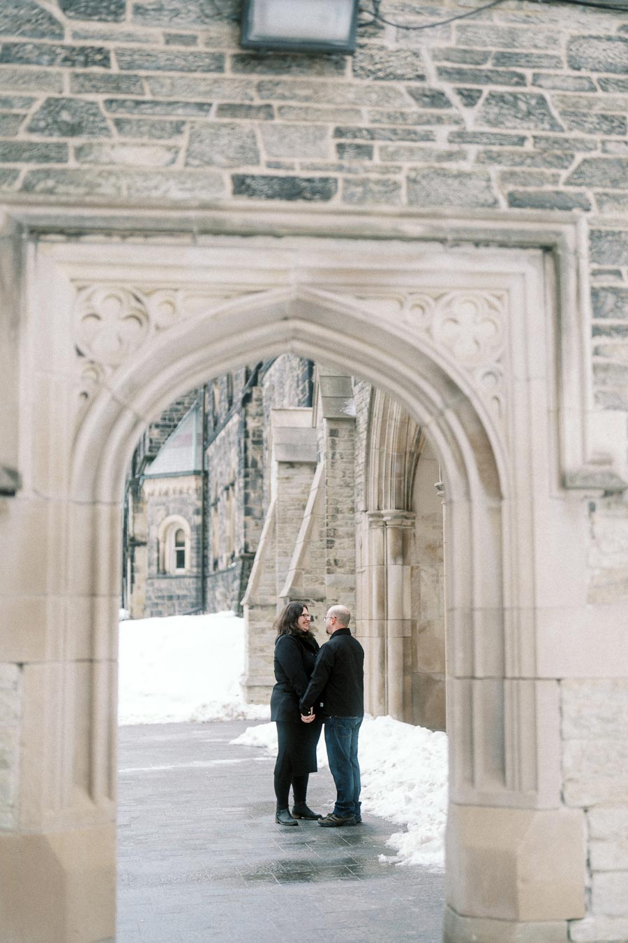 A couple holding hands under a historic stone archway on a snowy day, with a backdrop of an old building.
