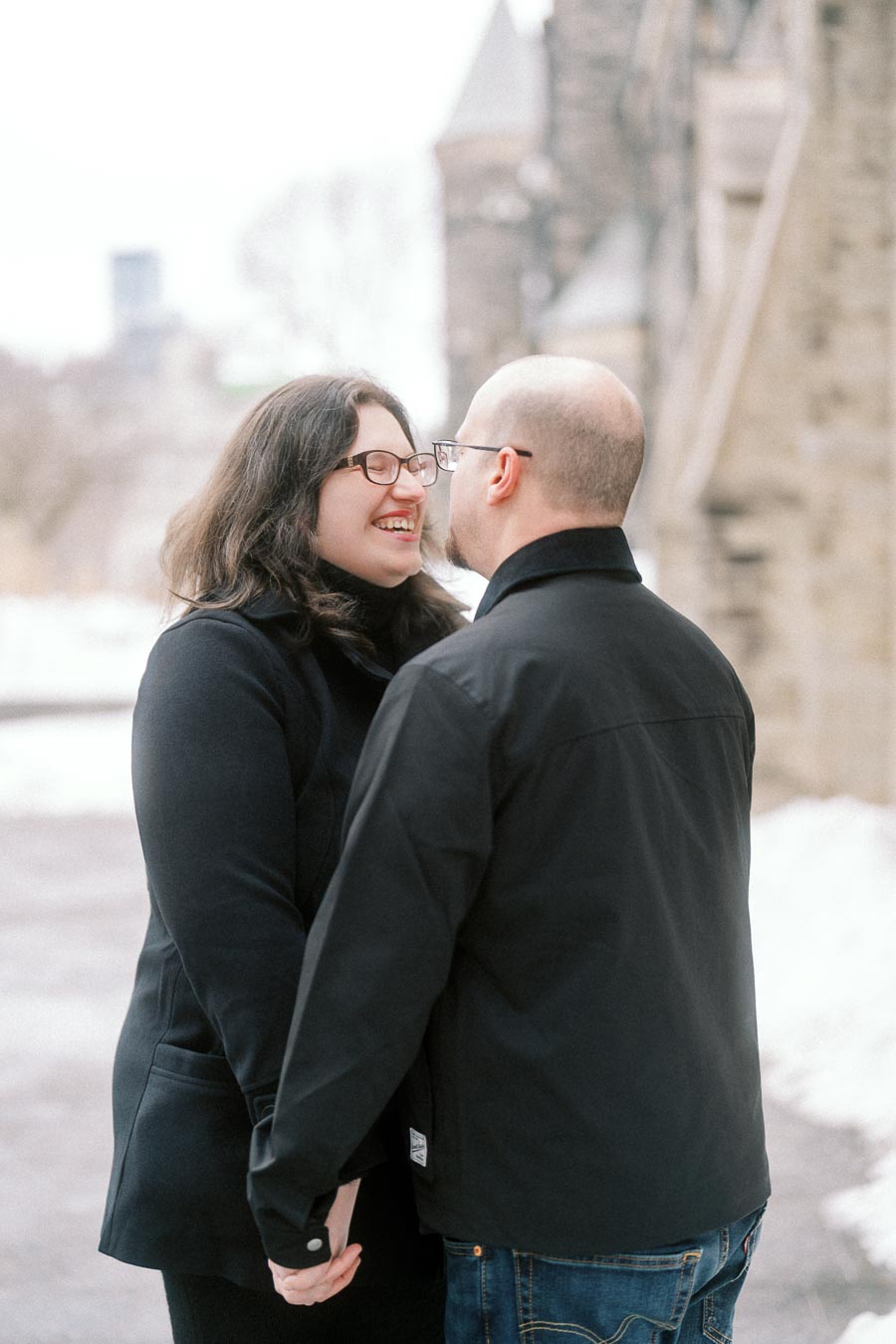 A couple sharing a joyful moment outdoors in a snowy setting, both wearing black jackets and glasses, standing close and smiling at each other near an old stone building.