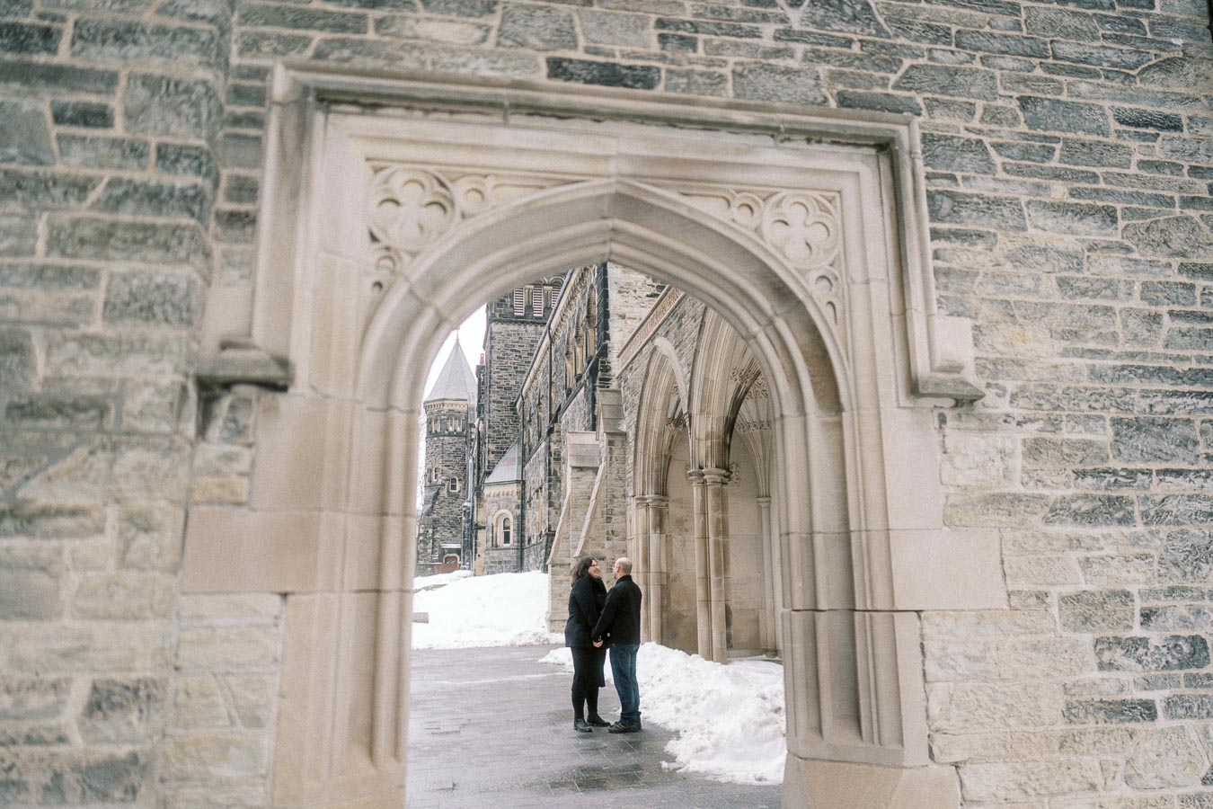 A couple holding hands stands under a stone archway, framed by historic architecture and surrounding snow.