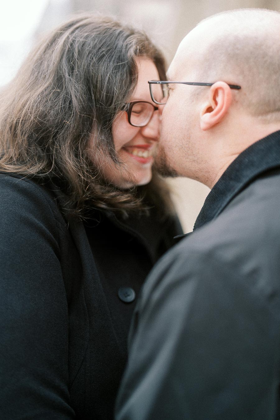 A couple sharing a loving moment, both wearing glasses and smiling as they lean in for a kiss, dressed in dark winter coats.