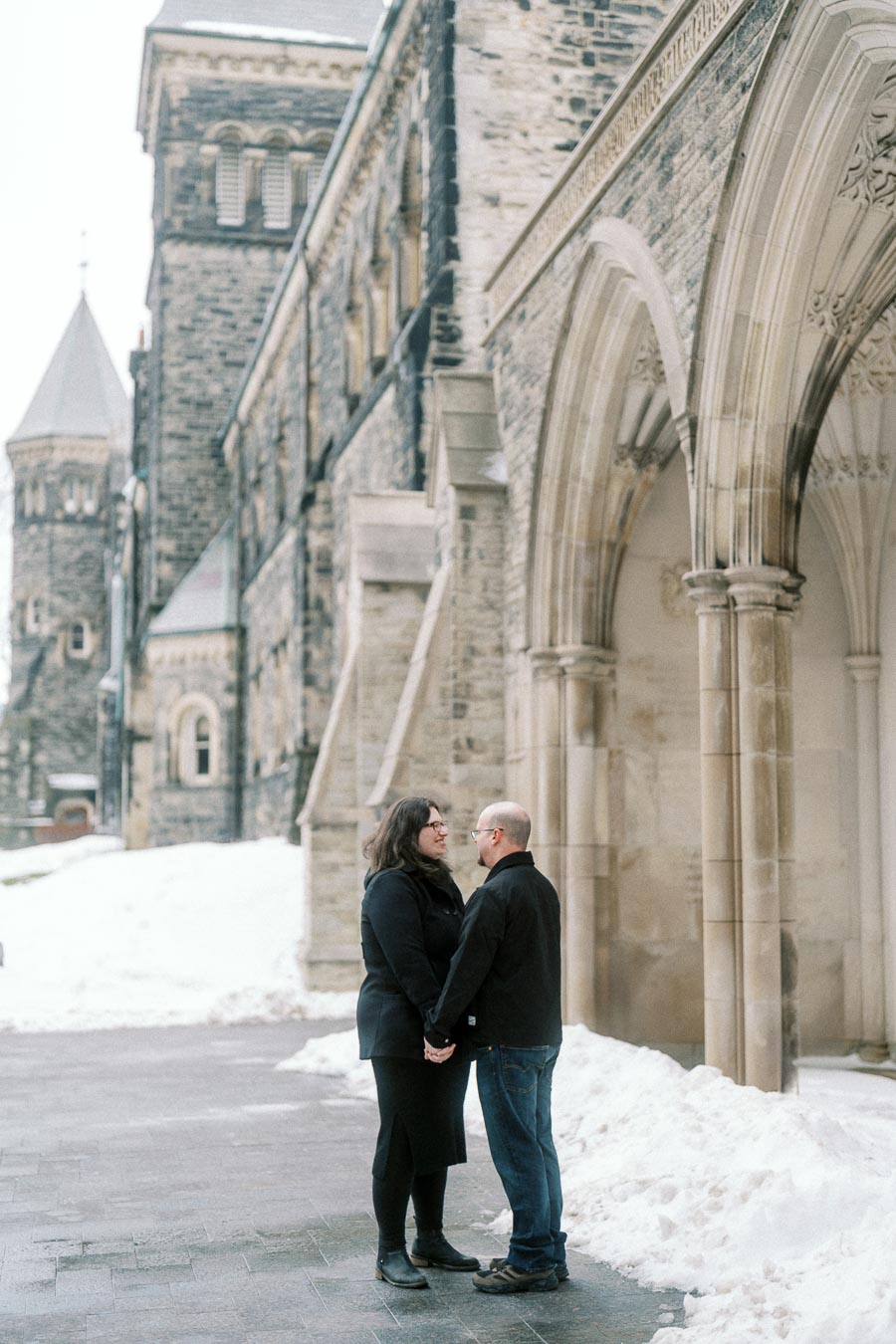 A couple holding hands and gazing at each other in front of a historic, stone building in a snowy landscape.